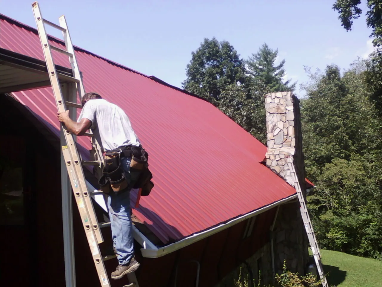 Roofer on a ladder beside a red metal roof and stone chimney — Soffit & Fascia Repair by Roofing Brothers in Branch